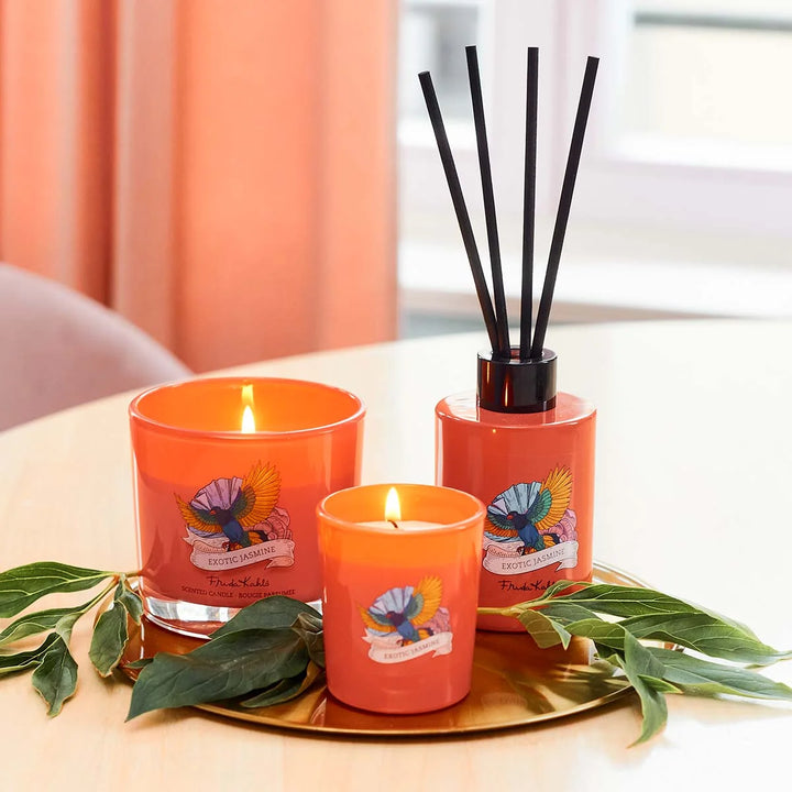 Frida Kahlo Exotic Jasmine scented candle. Bright orange glass candle featuring a colorful bird and floral design with elegant branding on the front.