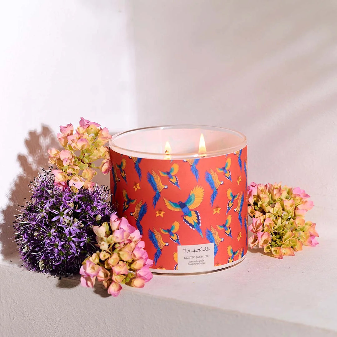 Frida Kahlo “Exotic Jasmine” scented candle with rose-gold lid and vibrant red label featuring colorful parrots and yellow flowers. 