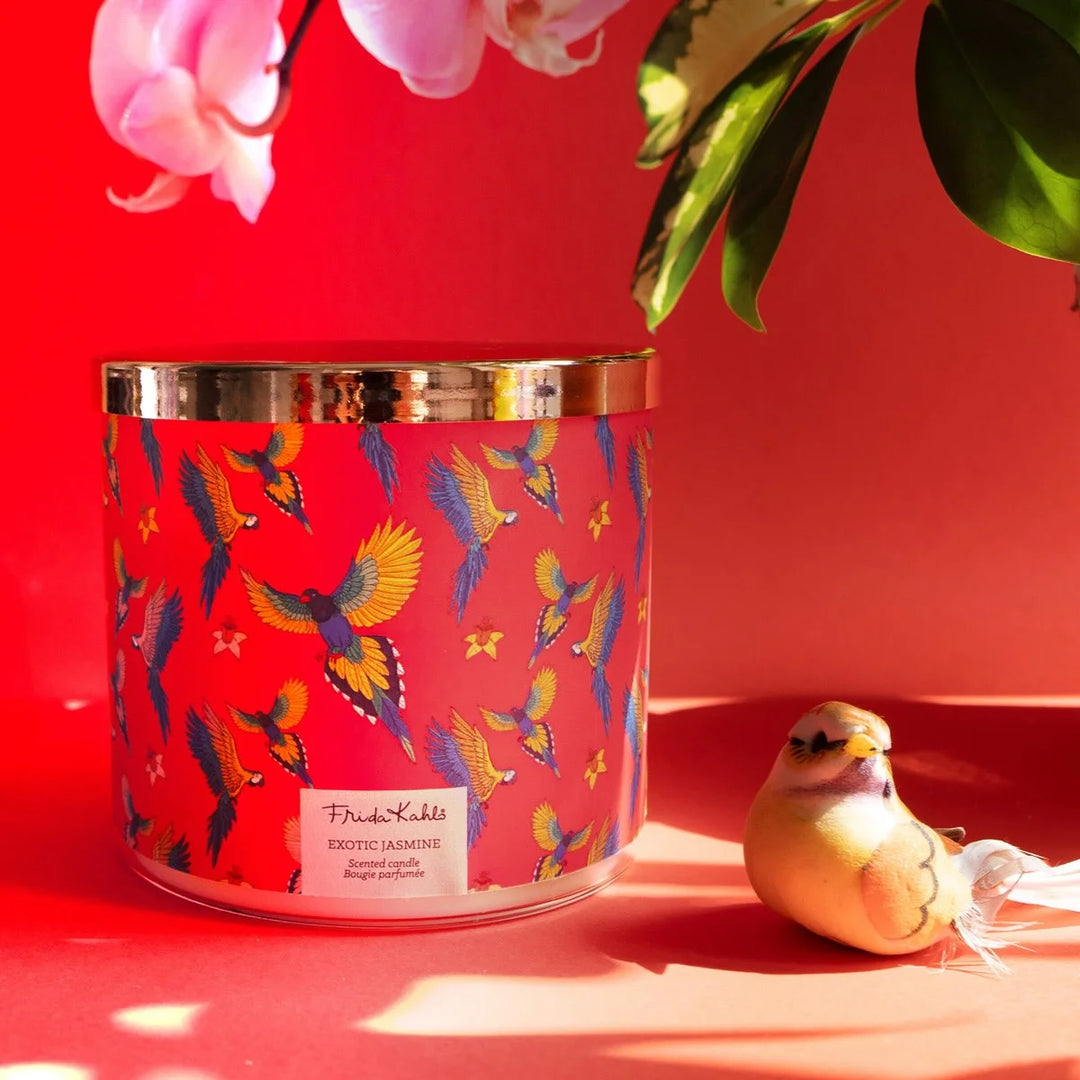 Frida Kahlo “Exotic Jasmine” scented candle with rose-gold lid and vibrant red label featuring colorful parrots and yellow flowers. 