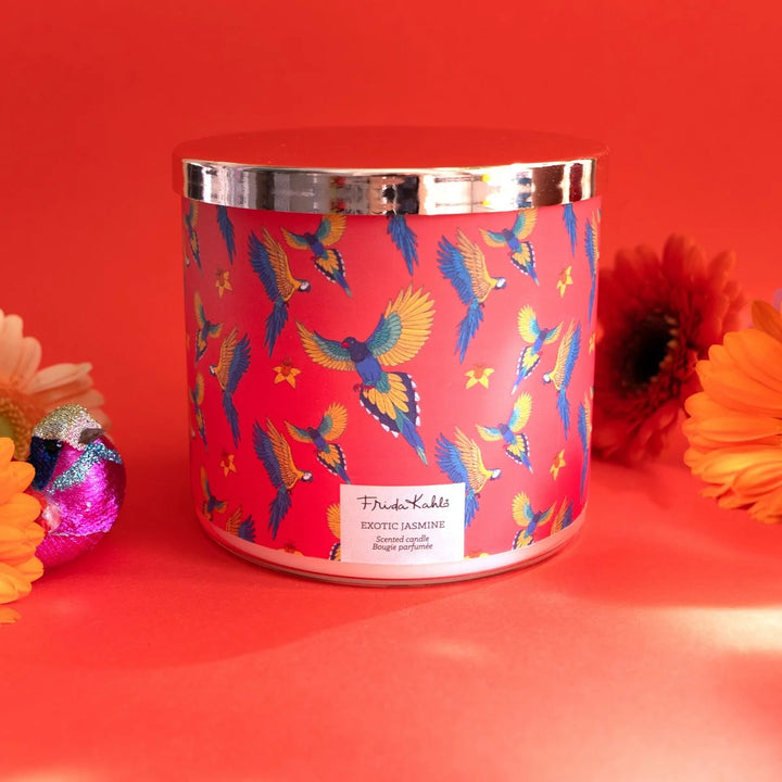 Frida Kahlo “Exotic Jasmine” scented candle with rose-gold lid and vibrant red label featuring colorful parrots and yellow flowers. 
