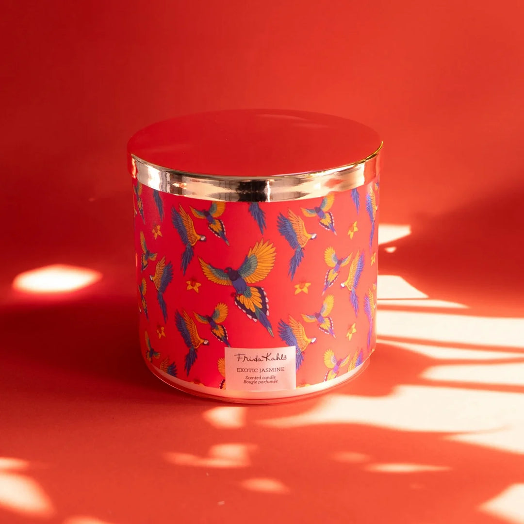 Frida Kahlo “Exotic Jasmine” scented candle with rose-gold lid and vibrant red label featuring colorful parrots and yellow flowers. 