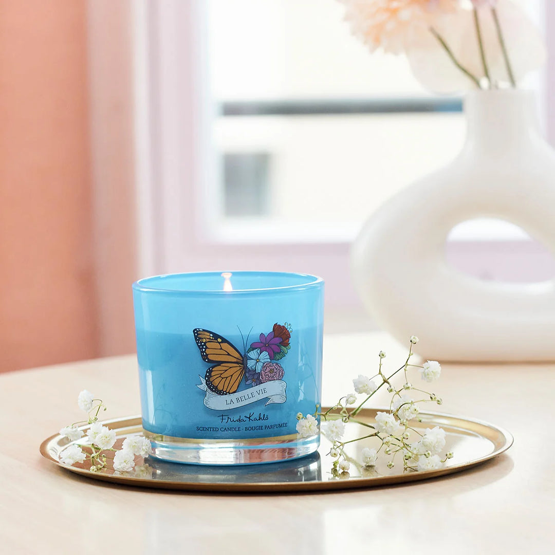 Frida Kahlo La Belle Vie scented candle. Blue glass candle featuring a vibrant butterfly and floral design with elegant Frida Kahlo branding on the front.