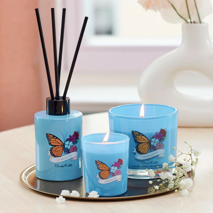 Frida Kahlo La Belle Vie scented candle. Blue glass candle featuring a vibrant butterfly and floral design with elegant Frida Kahlo branding on the front.