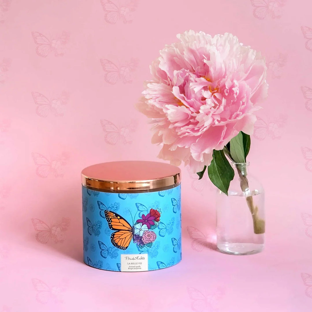 Frida Kahlo La Belle Vie scented candle. Large decorative candle with floral and butterfly design on a blue background and a metallic rose gold lid.