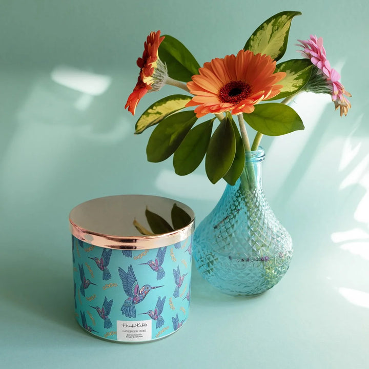 Frida Kahlo “Lavender Luxe” scented candle with a rose-gold lid and turquoise label decorated with colorful hummingbirds. 